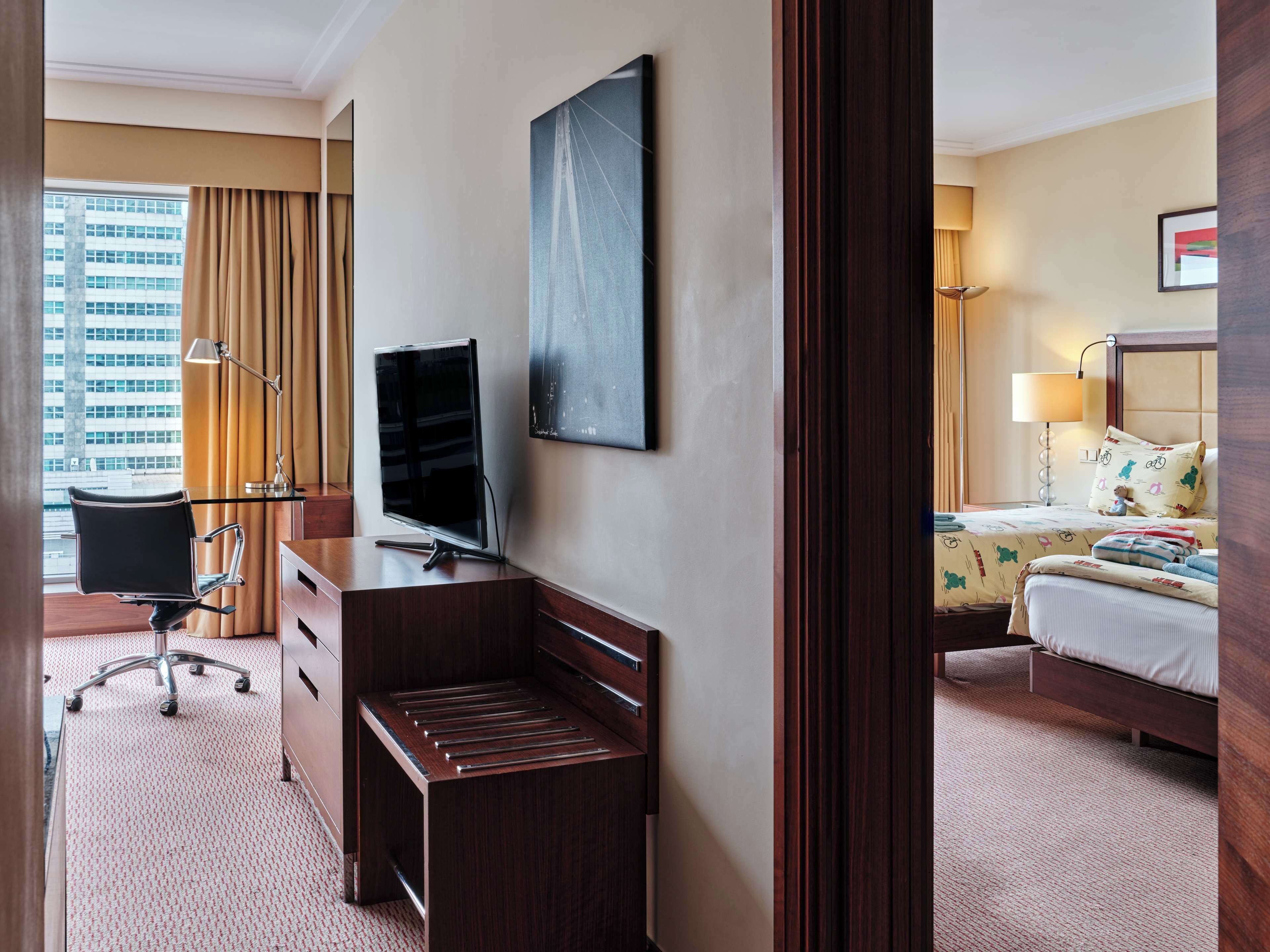 A hotel room with a desk, chair, television, bed, and a view of a building outside.
