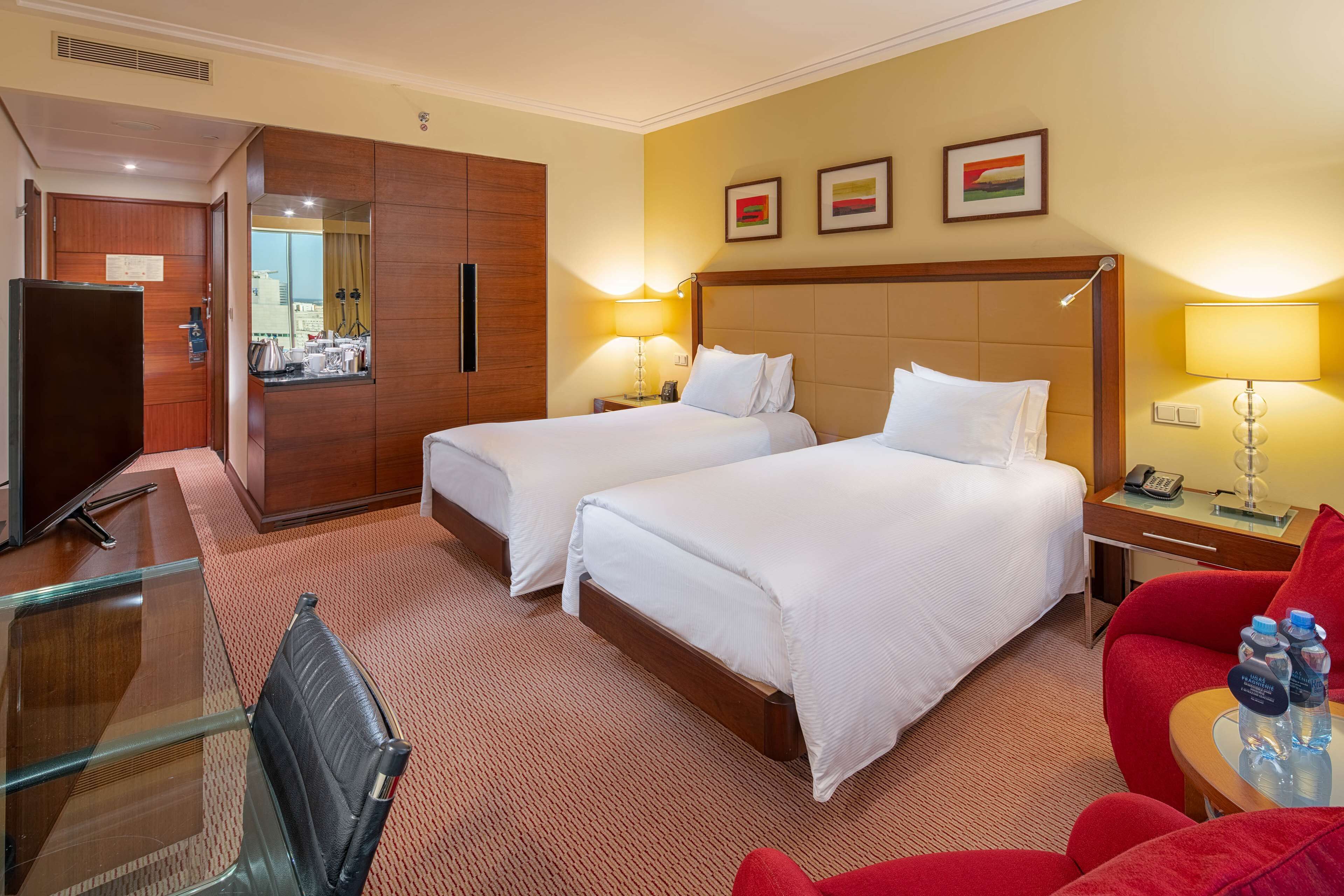 Een hotelkamer met twee bedden, een bureau met een stoel, een televisie en een rode fauteuil.