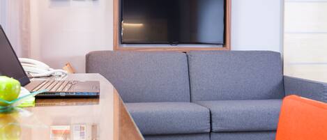 A modern room with a grey sofa, a wooden desk with a laptop, and a flat-screen TV mounted on the wall.