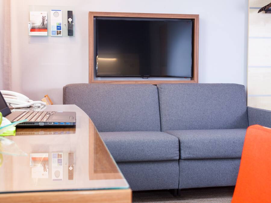 A modern room with a grey sofa, a wooden desk with a laptop, and a flat-screen TV mounted on the wall.