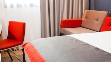 A hotel room with a bed, two red chairs, and a view of a cityscape through sheer curtains.