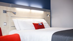 Une chambre d’hôtel avec un lit bien fait, une chaise rouge et une lampe fixée au mur.