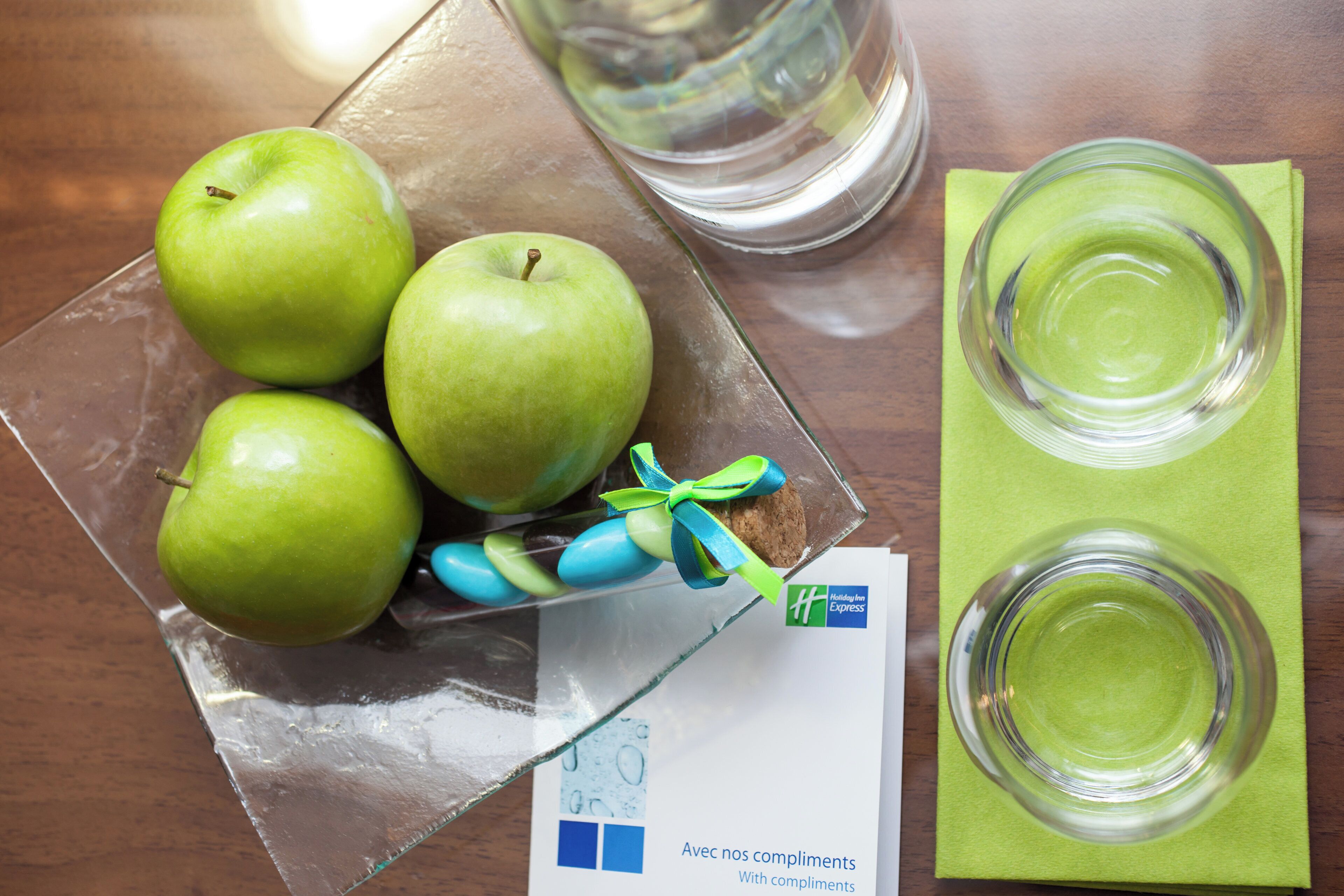 Een glazen schaal met groene appels, een koekje en een kaartje op een tafel.