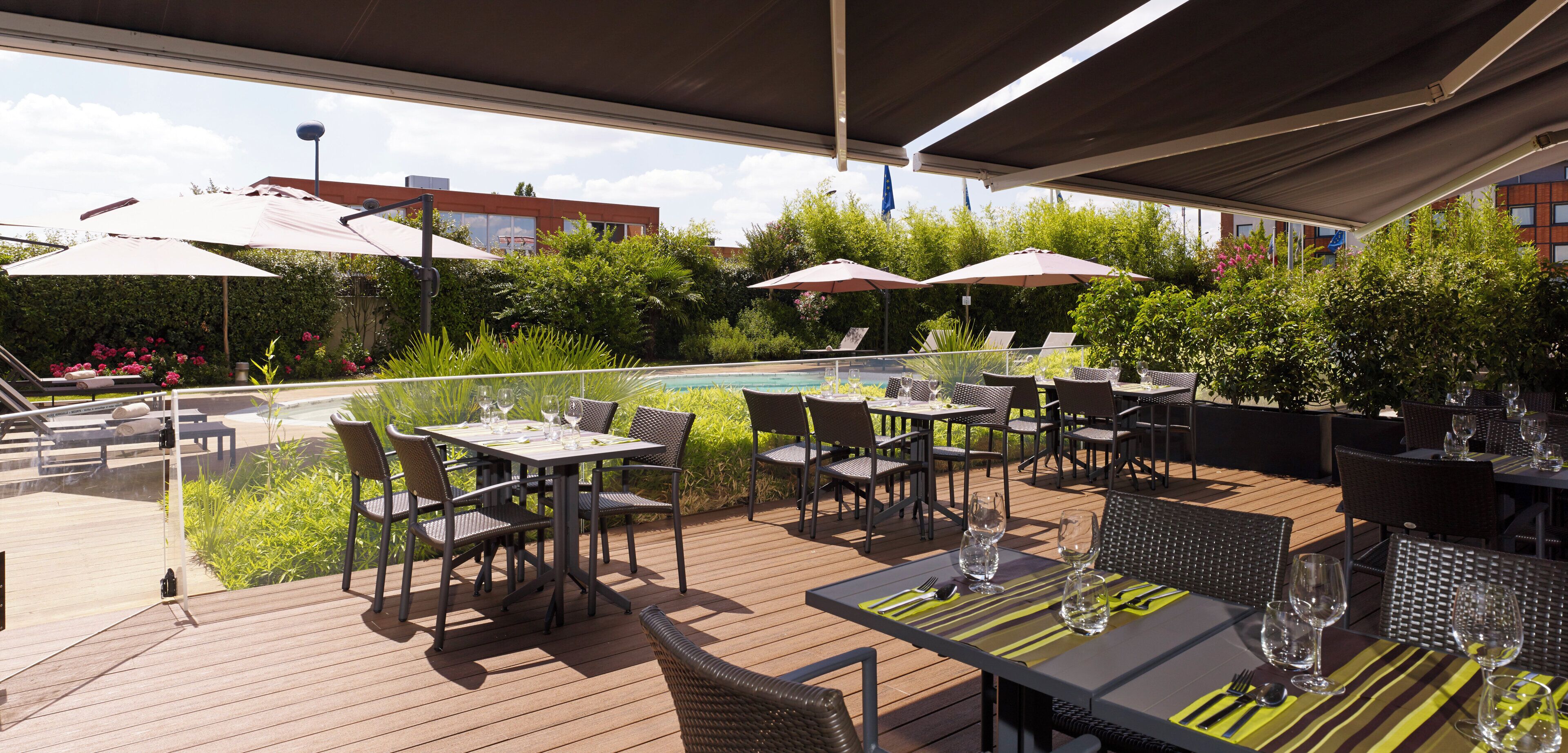 An outdoor dining area with tables and chairs, umbrellas, and a pool in the background.