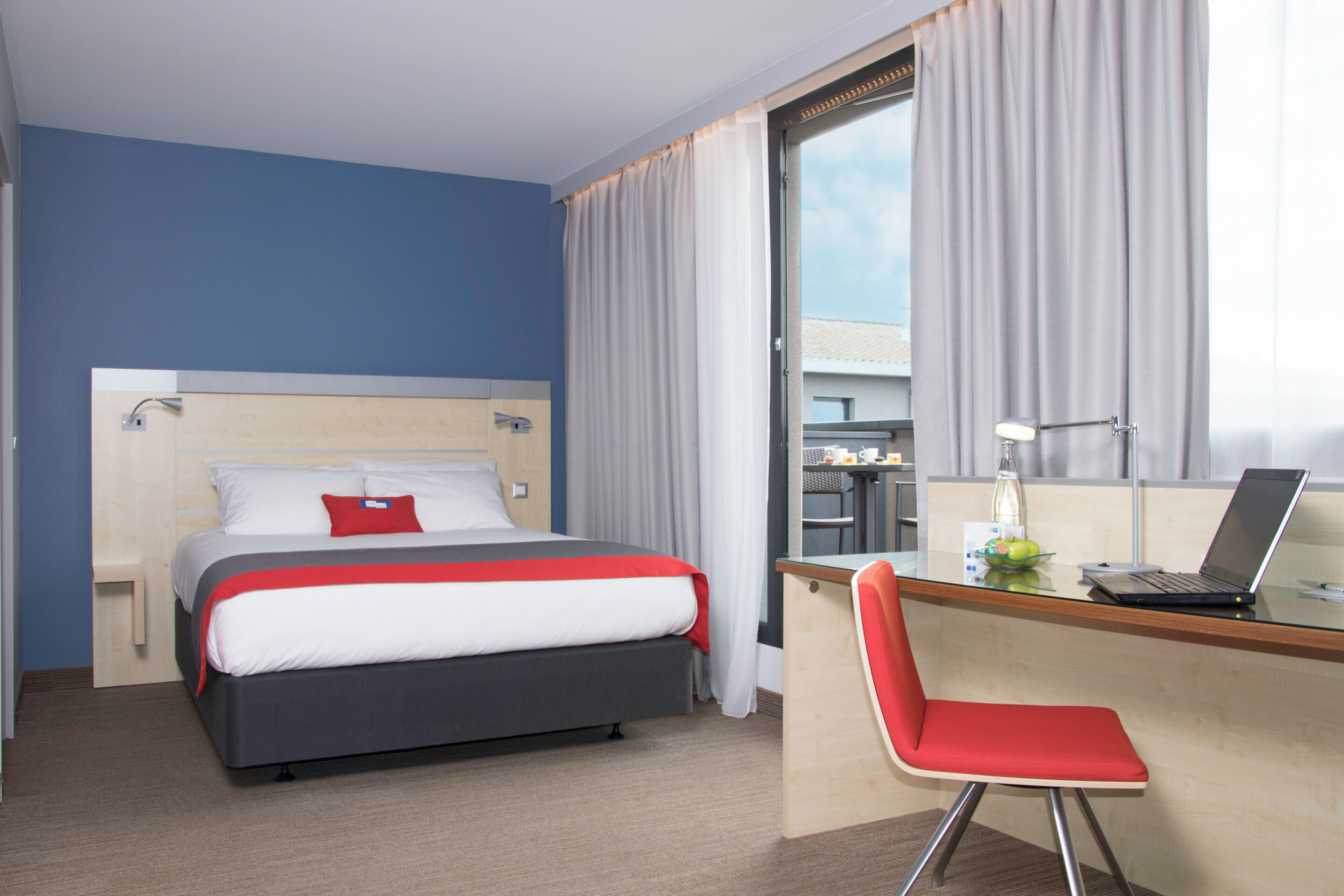 A hotel room with a bed, a desk with a laptop, a chair, and a window with curtains.