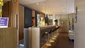 A modern hotel lobby with a reception desk, bar area with stools, and a seating area with a sofa and chairs. - Holiday Inn Express Toulouse Airport by IHG (Blagnac)