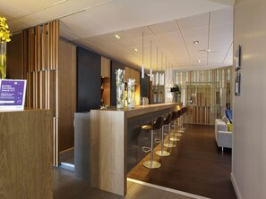 Eine moderne Hotelhalle mit Empfangstresen, Barbereich mit Hockern und einer Sitzecke mit Sofa und Sesseln.