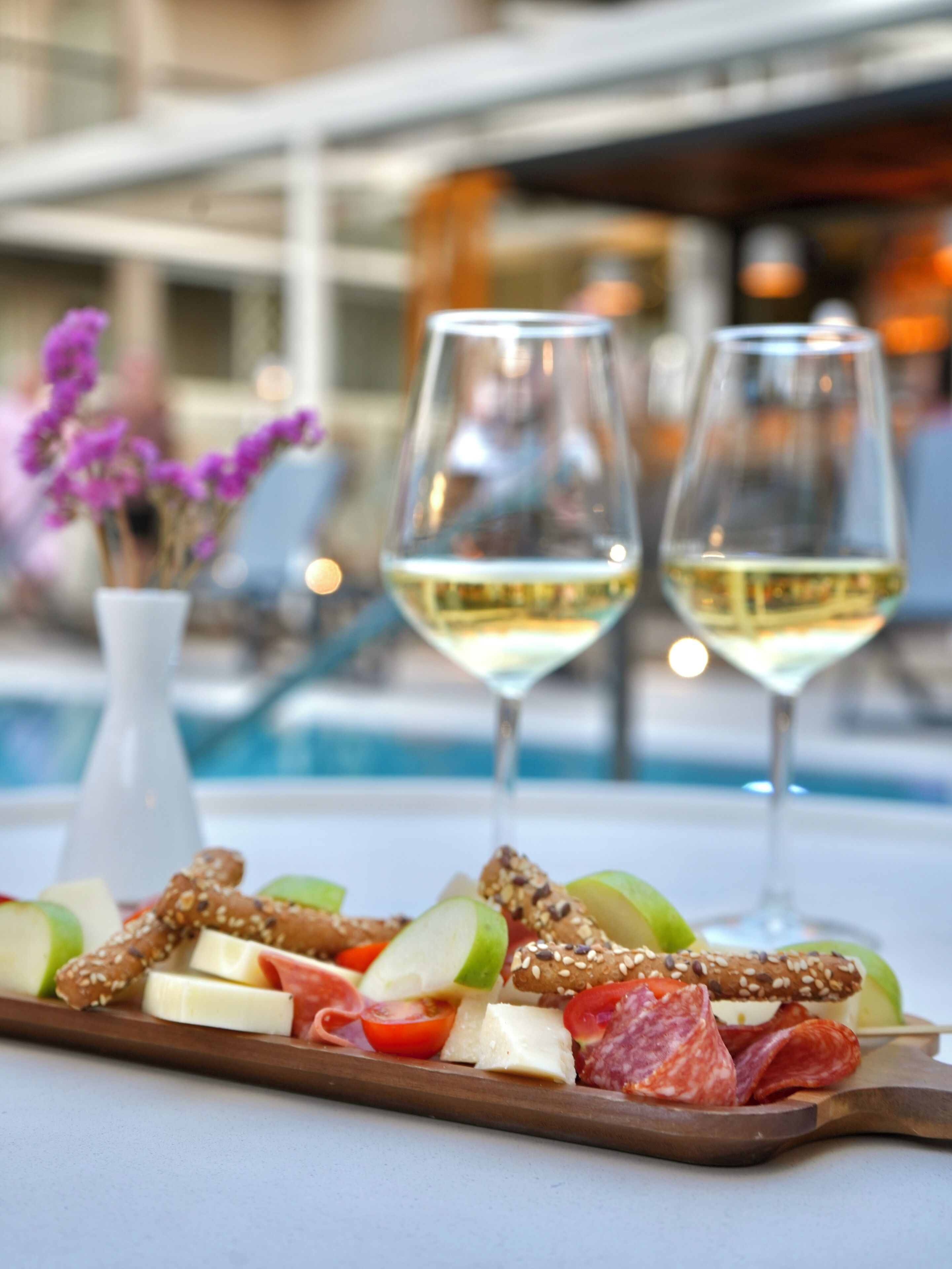 A tray of appetizers including cheese, nuts, and meats, with two glasses of white wine, set on a table by a poolside.