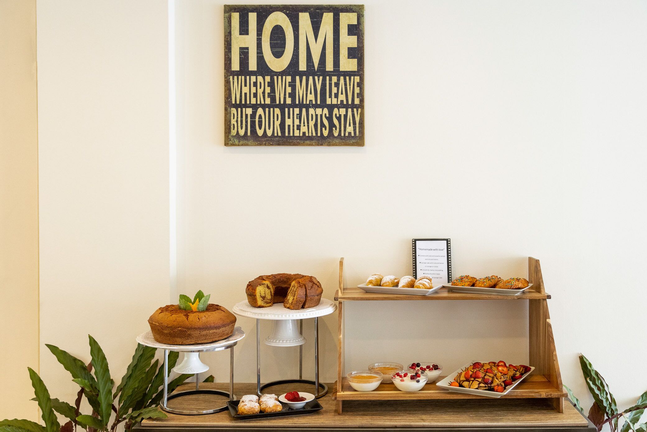 A dessert display with cakes and pastries, a sign above stating "HOME WHERE WE MAY LEAVE BUT OUR HEARTS STAY", and a wooden shelf with more pastries.
