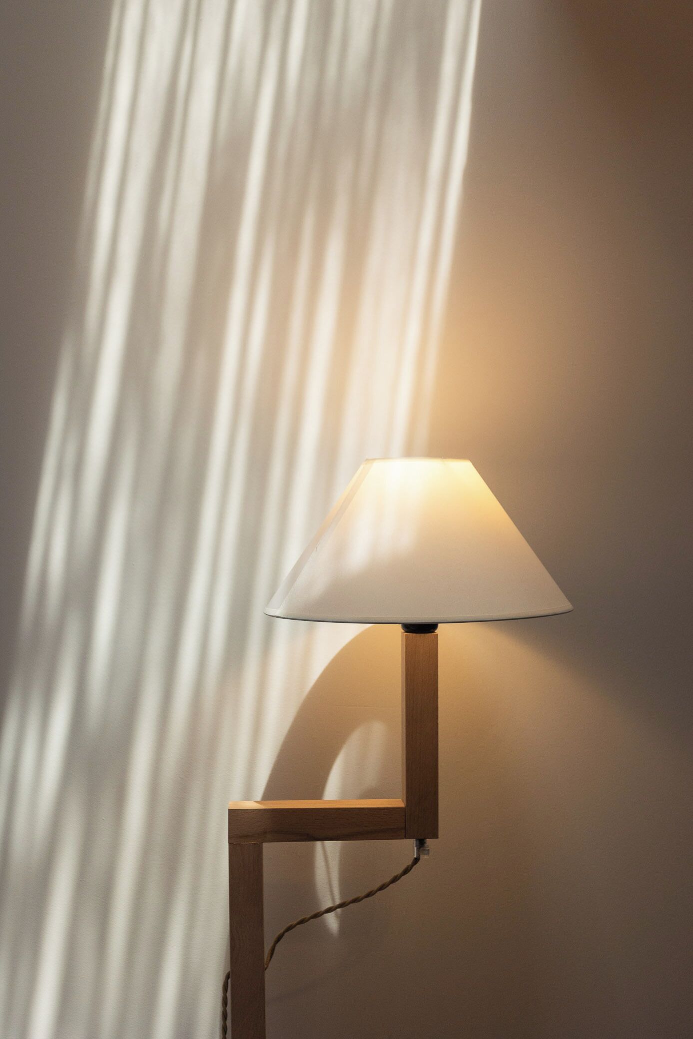 A lamp with a white shade and a wooden base, positioned against a wall with vertical blinds.