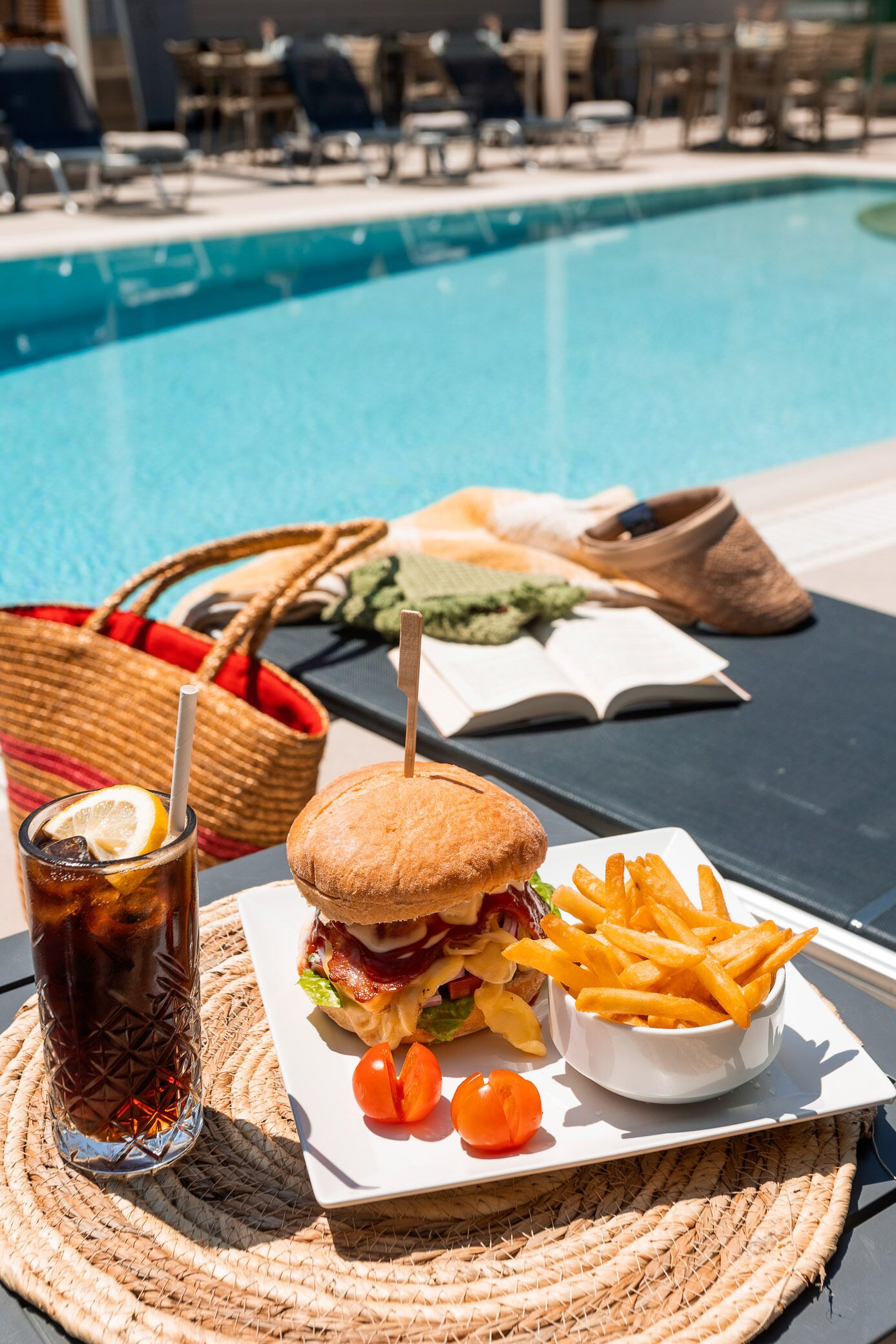 A poolside dining setup with a sandwich, fries, and a drink on a woven mat.