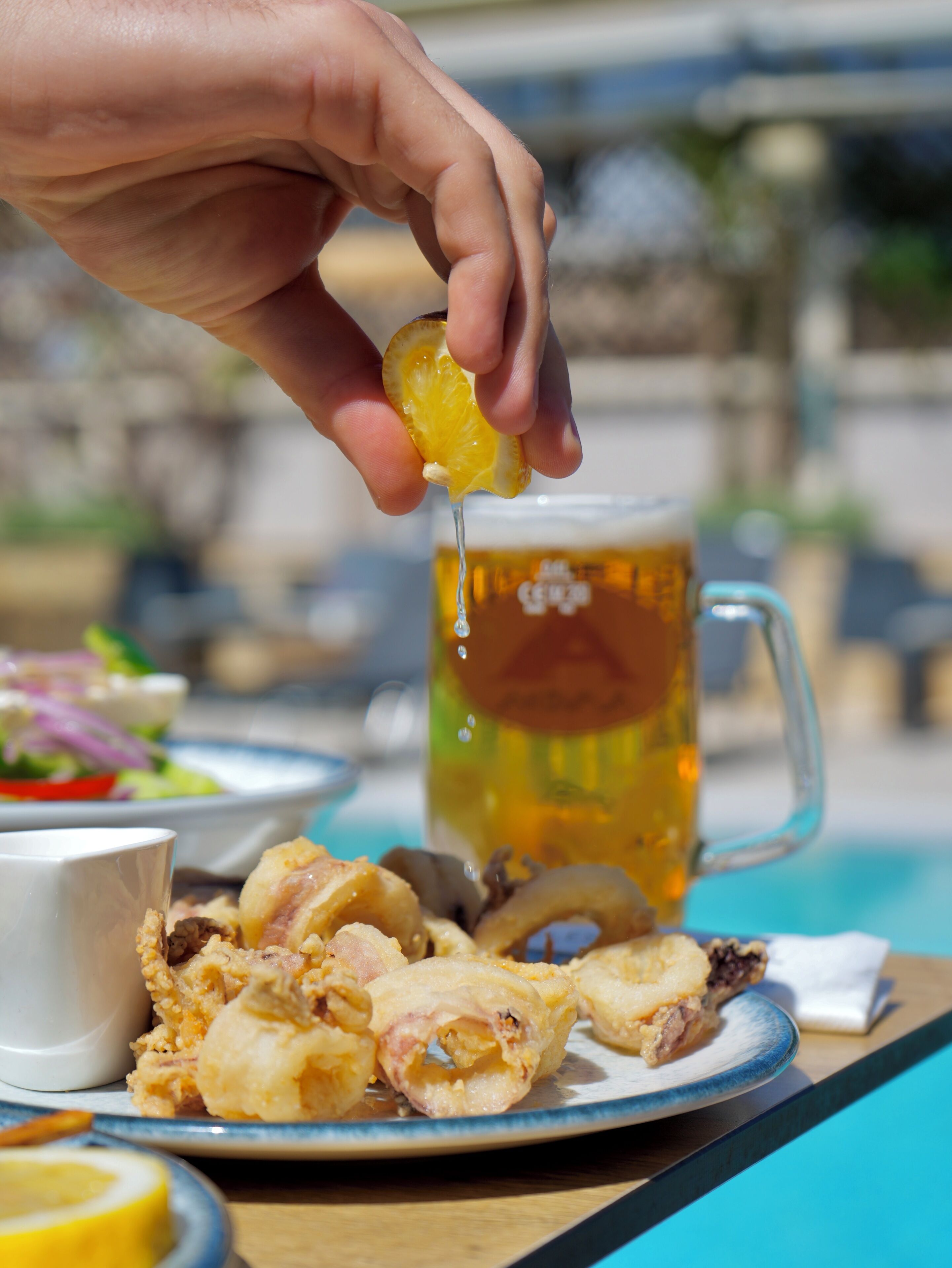 A hand squeezing lemon over a plate of fried food, with a beer mug and a cup in the background.