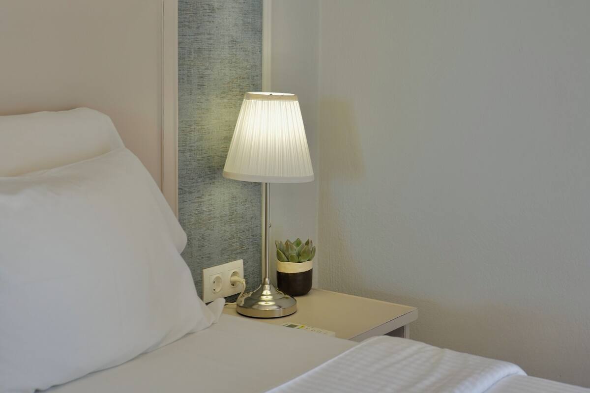 A neatly made bed with white linens, a bedside lamp, and a small potted plant on a bedside table.