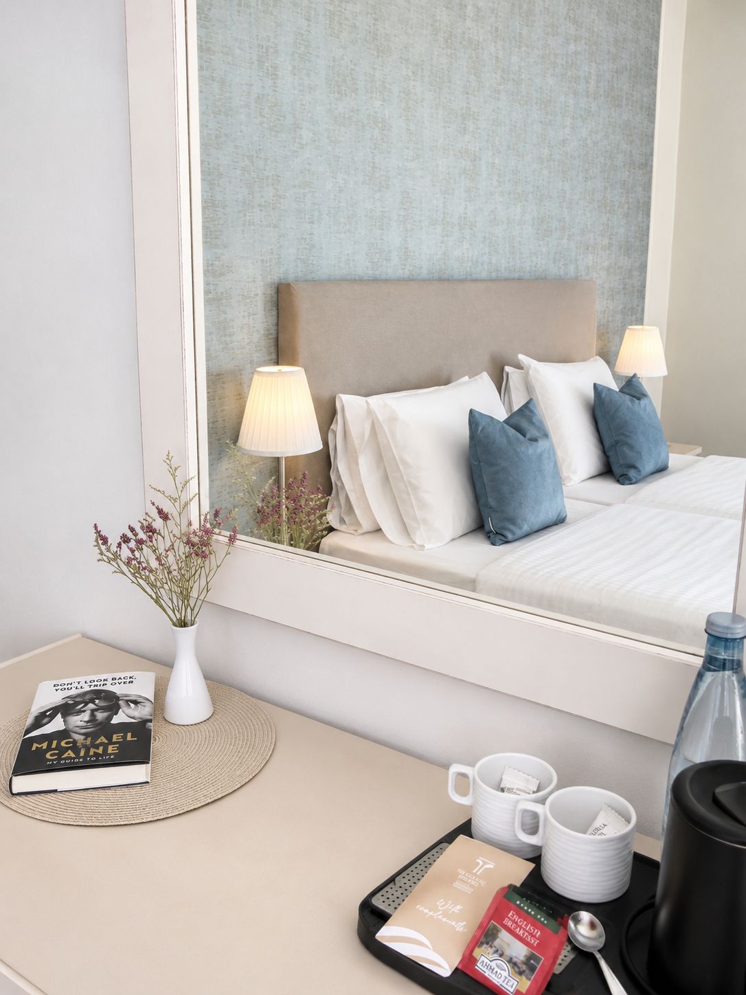 A hotel room with a bed, bedside lamps, a bedside table with a book, a vase with flowers, and a tray with breakfast items.