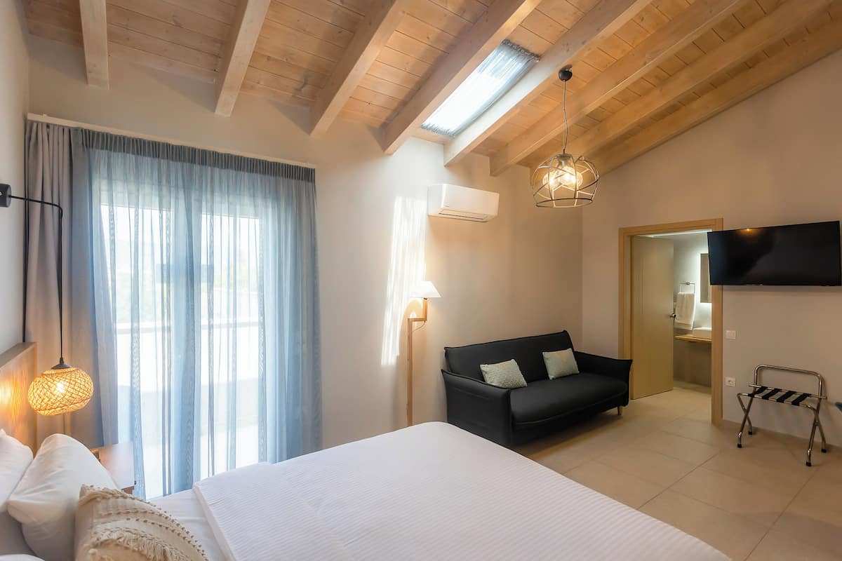 A hotel room with a bed, a sofa, a TV, a window with curtains, and a ceiling with wooden beams.