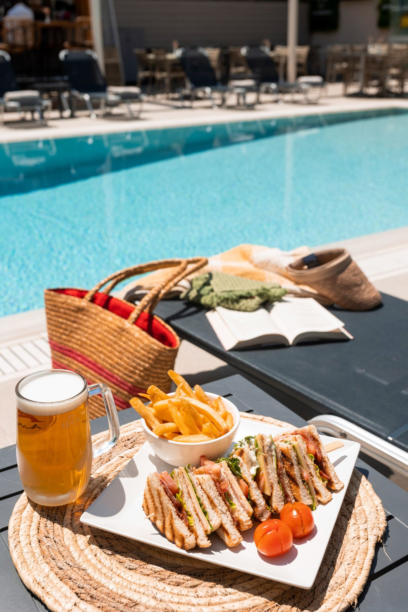 A poolside table with a plate of sandwiches, fries, and a beer mug.