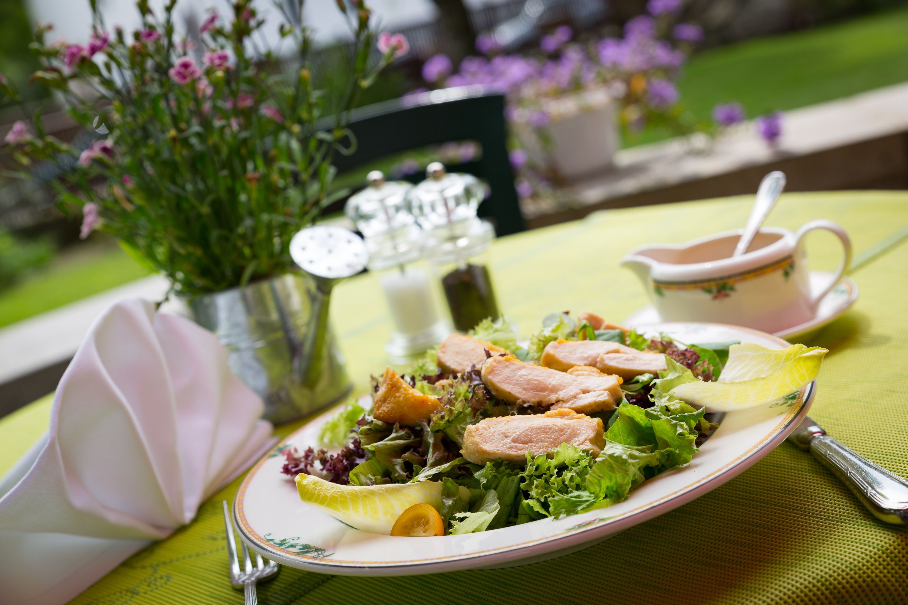 Ein Salat mit verschiedenen Blattsalaten und Fleischscheiben, eine Suppentasse und ein kleiner Krug auf einem Tisch mit grüner Tischdecke.