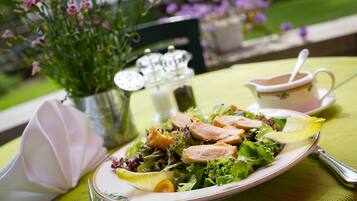 Ein Salat mit verschiedenen Blattsalaten und Fleischscheiben, eine Suppentasse und ein kleiner Krug auf einem Tisch mit grüner Tischdecke.