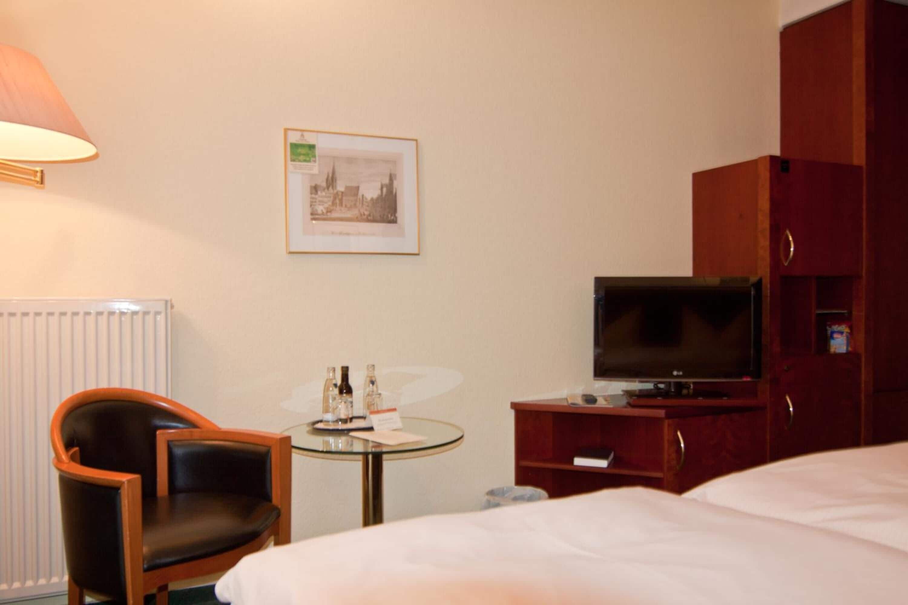 Una habitación de hotel con una cama, un televisor sobre un mueble de madera, una mesita con una silla y un cuadro en la pared.