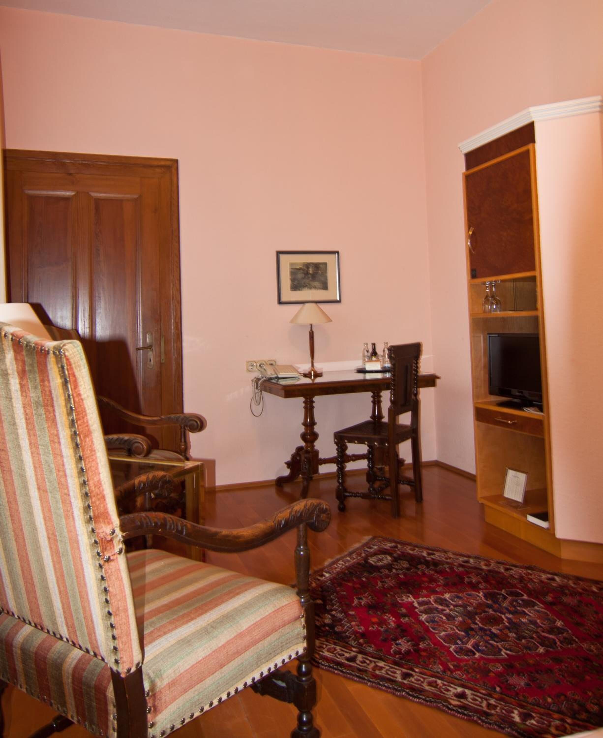 Ein Zimmer mit einem Holzstuhl, einem kleinen Tisch mit Lampe, einem Fernseher auf einer Holzkommode und einem Teppich mit Muster.