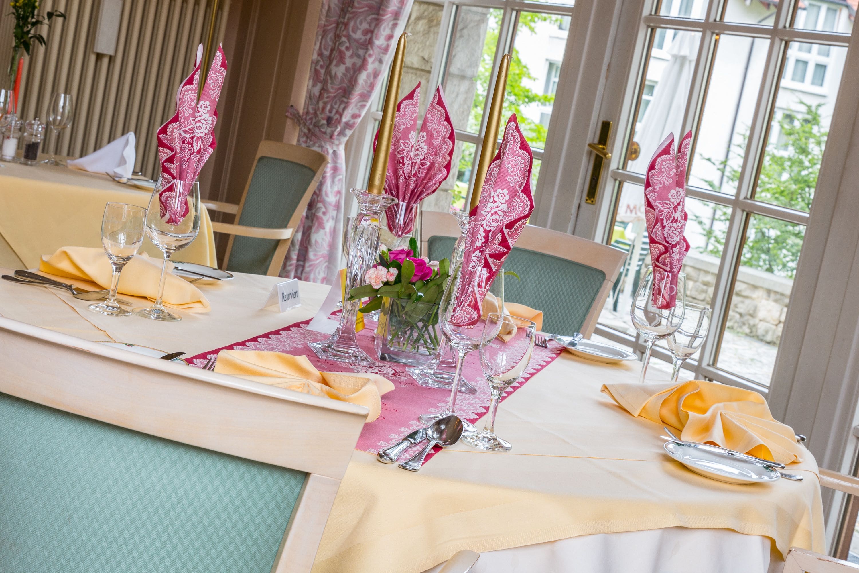Una mesa de comedor dispuesta con servilletas rosas, cristalería transparente y un jarrón con flores.