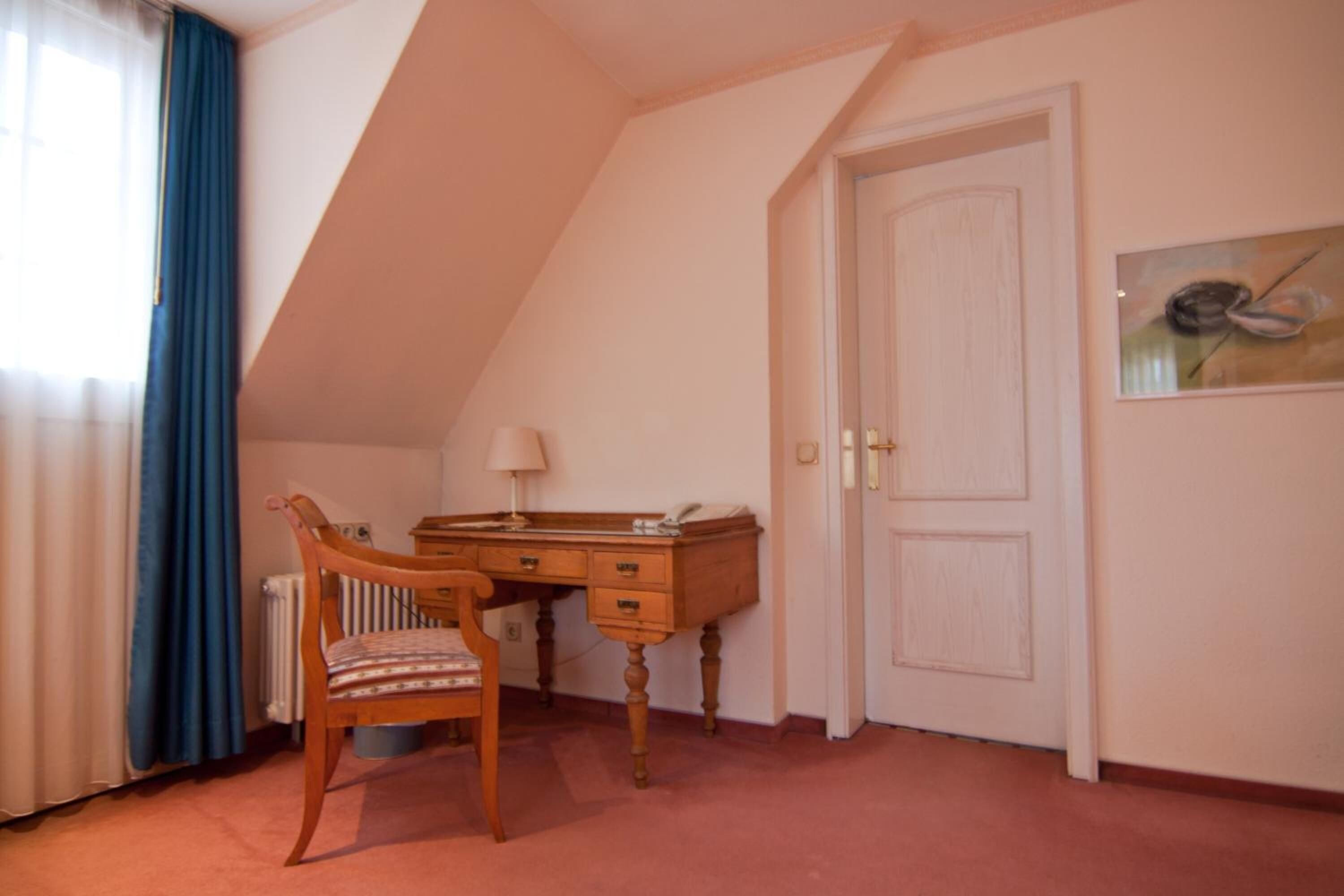 Una habitación con techo inclinado, un escritorio de madera con una silla, un radiador, una puerta y un cuadro en la pared.