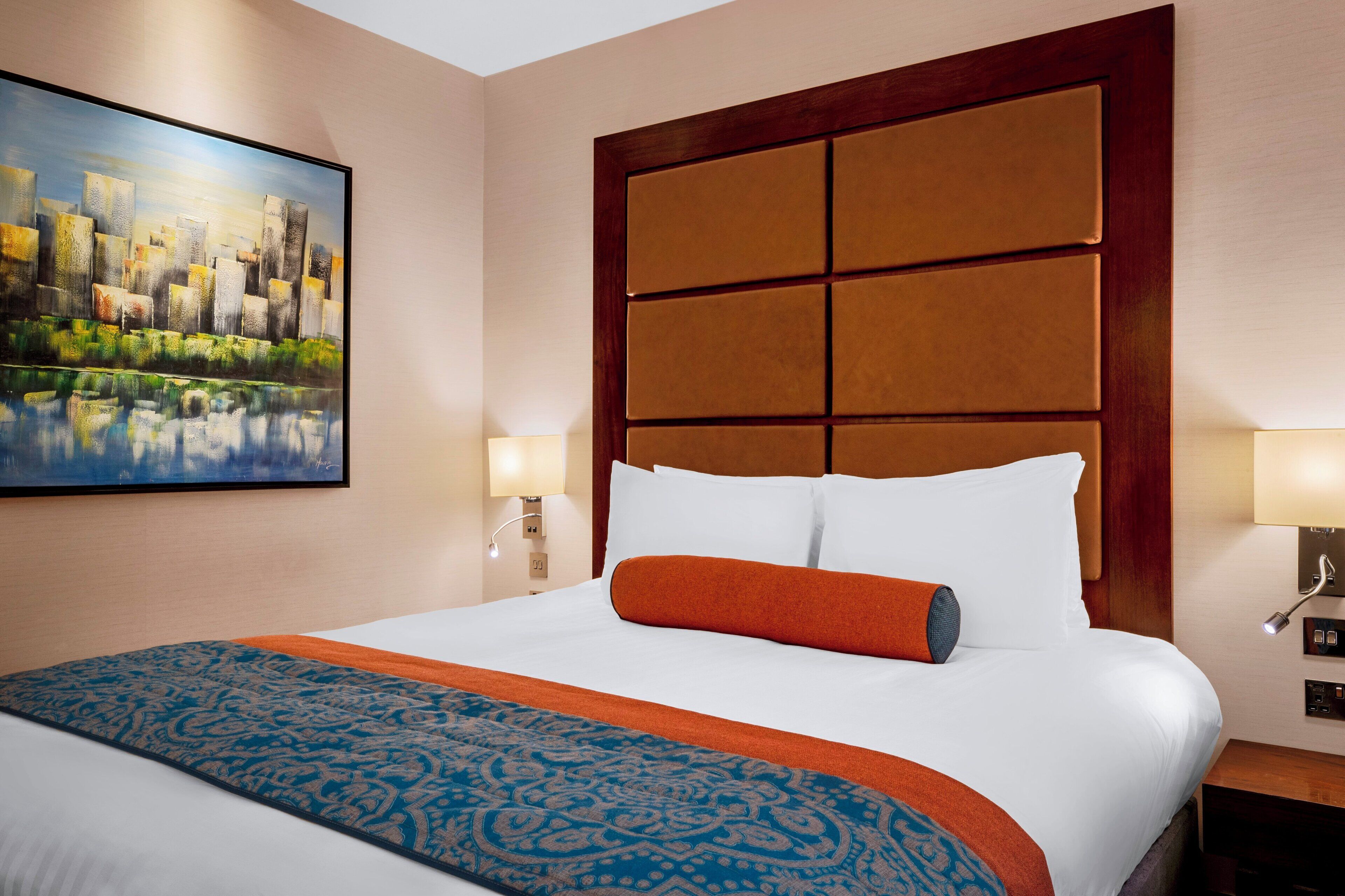 A hotel room with a large bed, a cityscape painting on the wall, and two bedside lamps.