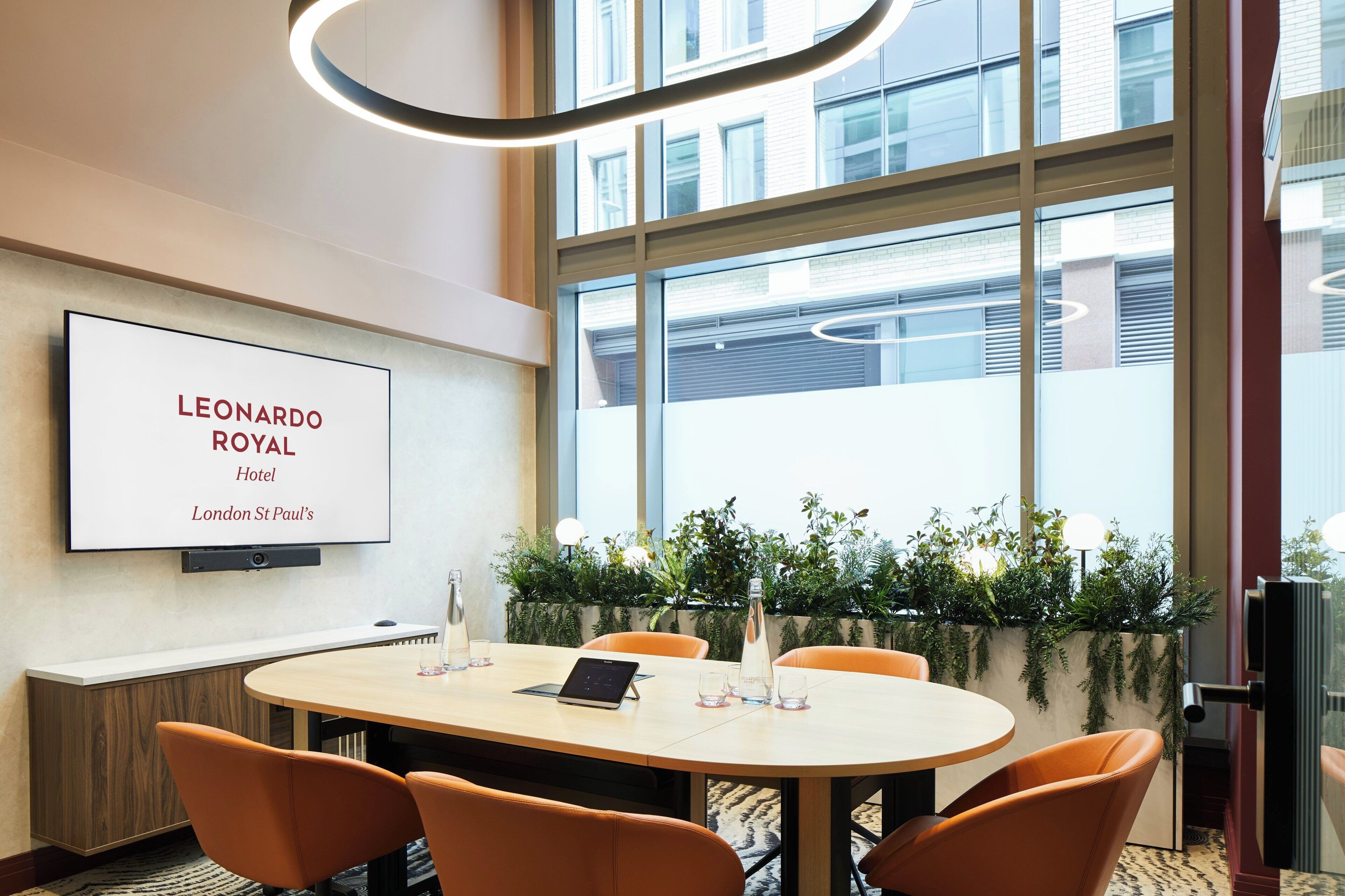 A conference room with a large table, four chairs, a mounted TV displaying 'LEONARDO ROYAL Hotel London St Paul's', and a view of the outside through large windows.