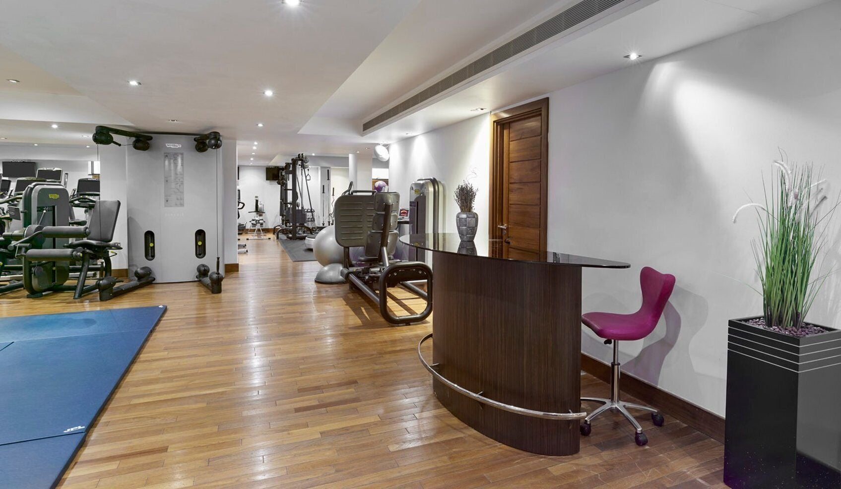 A gym with exercise equipment, a reception desk, and a reception area with a chair and a plant.