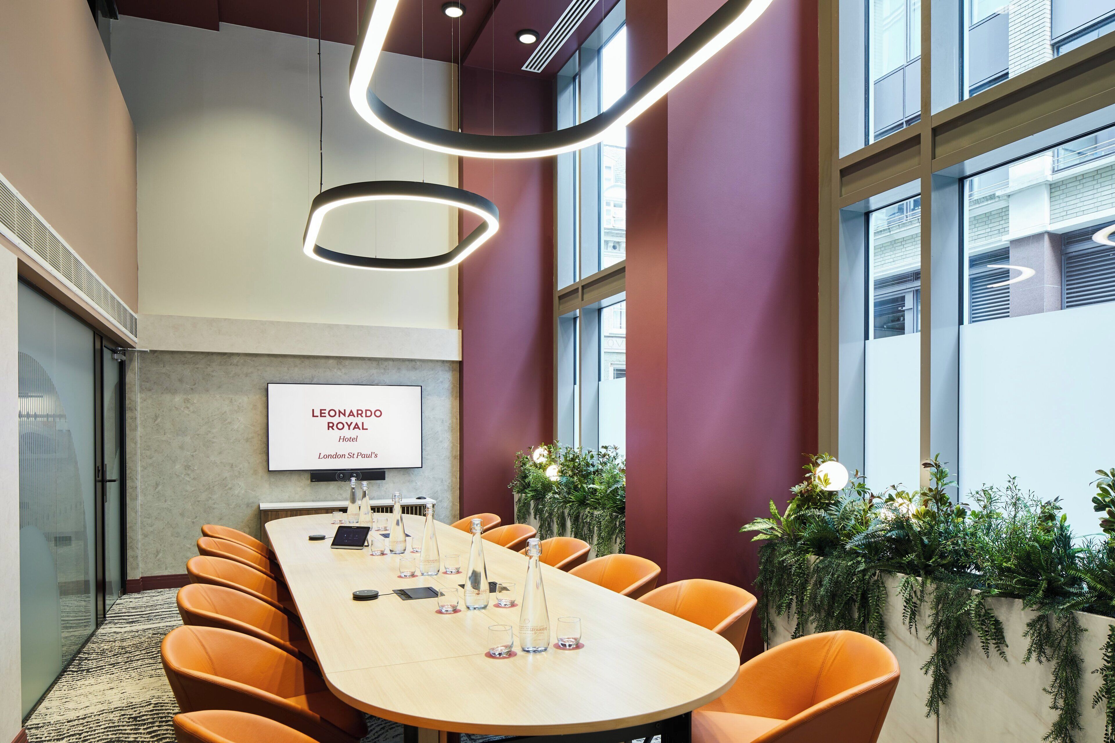 Ein Konferenzraum mit einem langen Tisch, orangefarbenen Stühlen, einem Bildschirm mit Hotelinformationen und großen Fenstern mit Blick auf Gebäude.