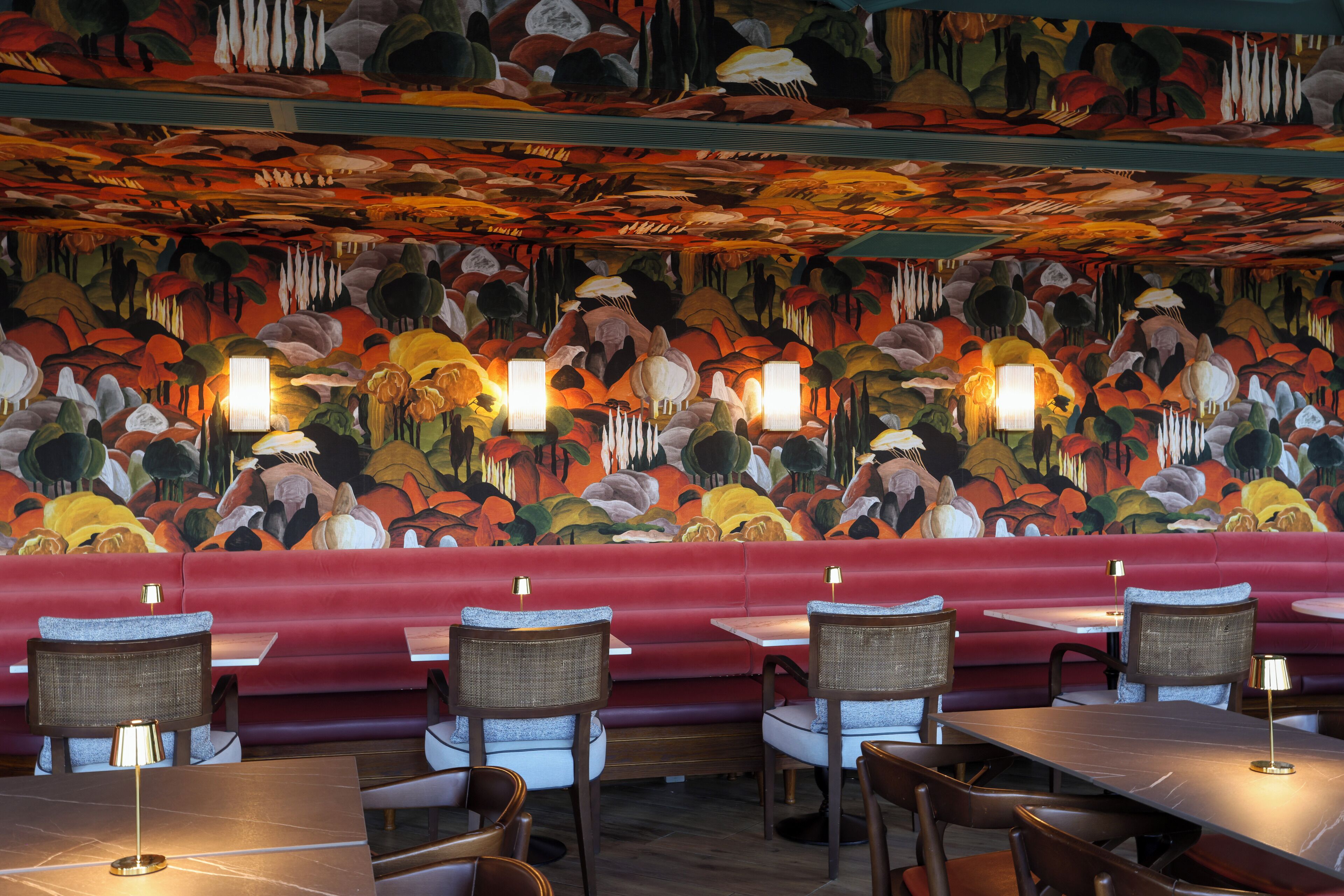 Interno di un ristorante con un murale vivace, sedute rosse e tavoli illuminati da lampade.