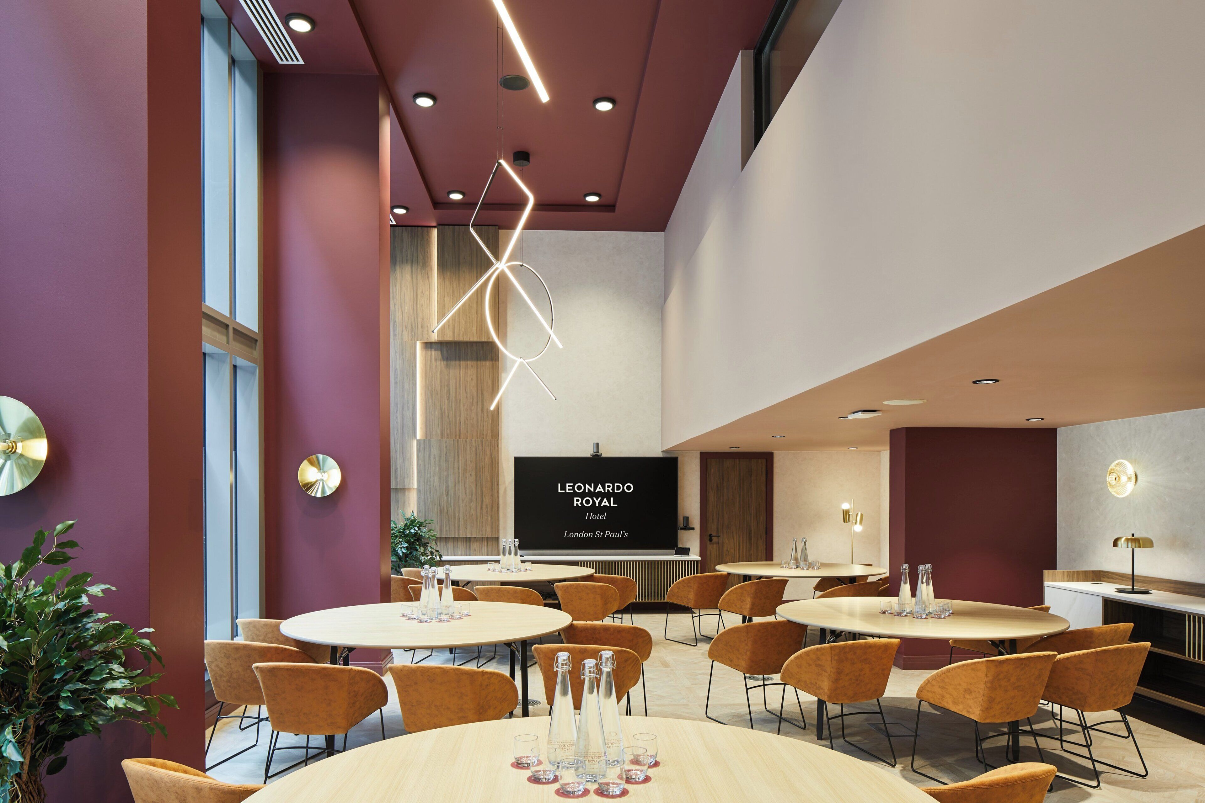 A modern hotel conference room with round tables, orange chairs, and a large screen displaying the hotel name.