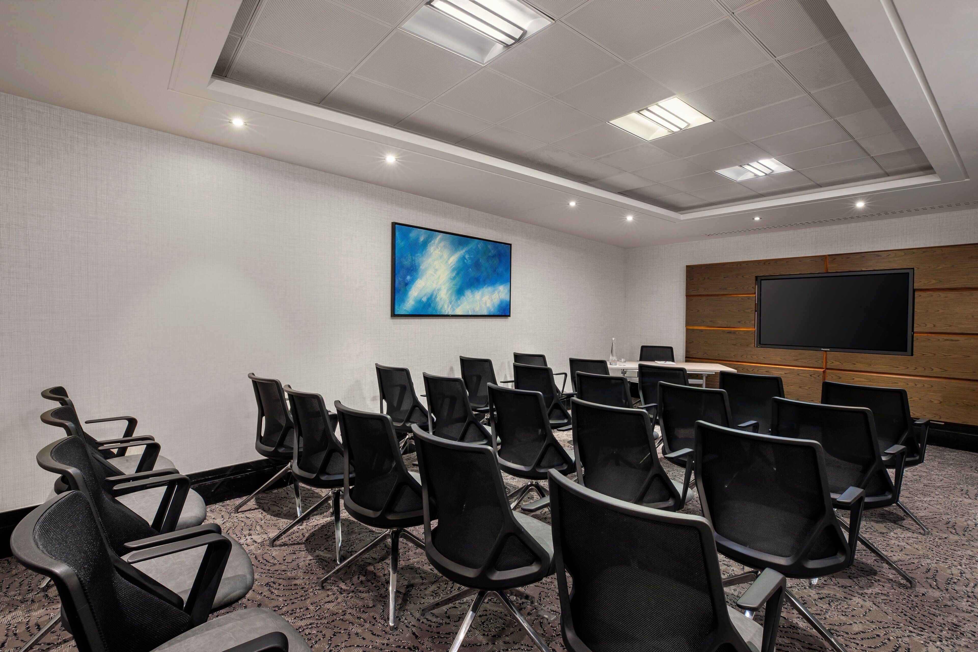 A conference room with rows of black chairs, a large screen, and a blue painting on the wall.
