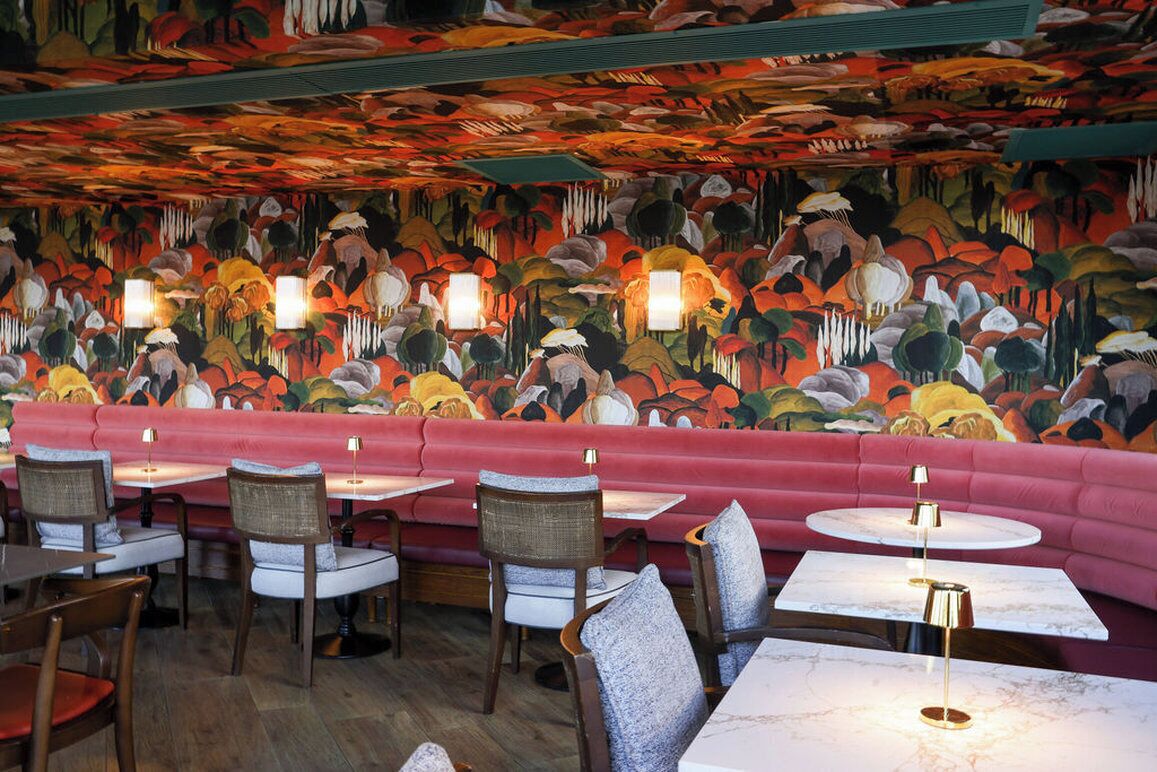 Interior de un restaurante con un mural llamativo, asientos acolchados y mesas redondas con lámparas.