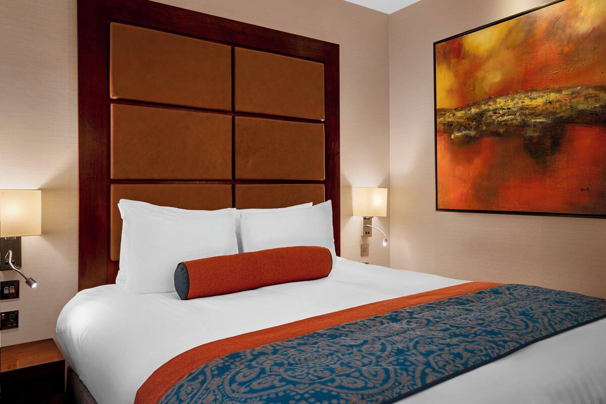 Ein Hotelzimmer mit einem großen Bett, zwei Nachttischlampen, einem Kopfteil mit Panelmuster und einem Gemälde an der Wand.