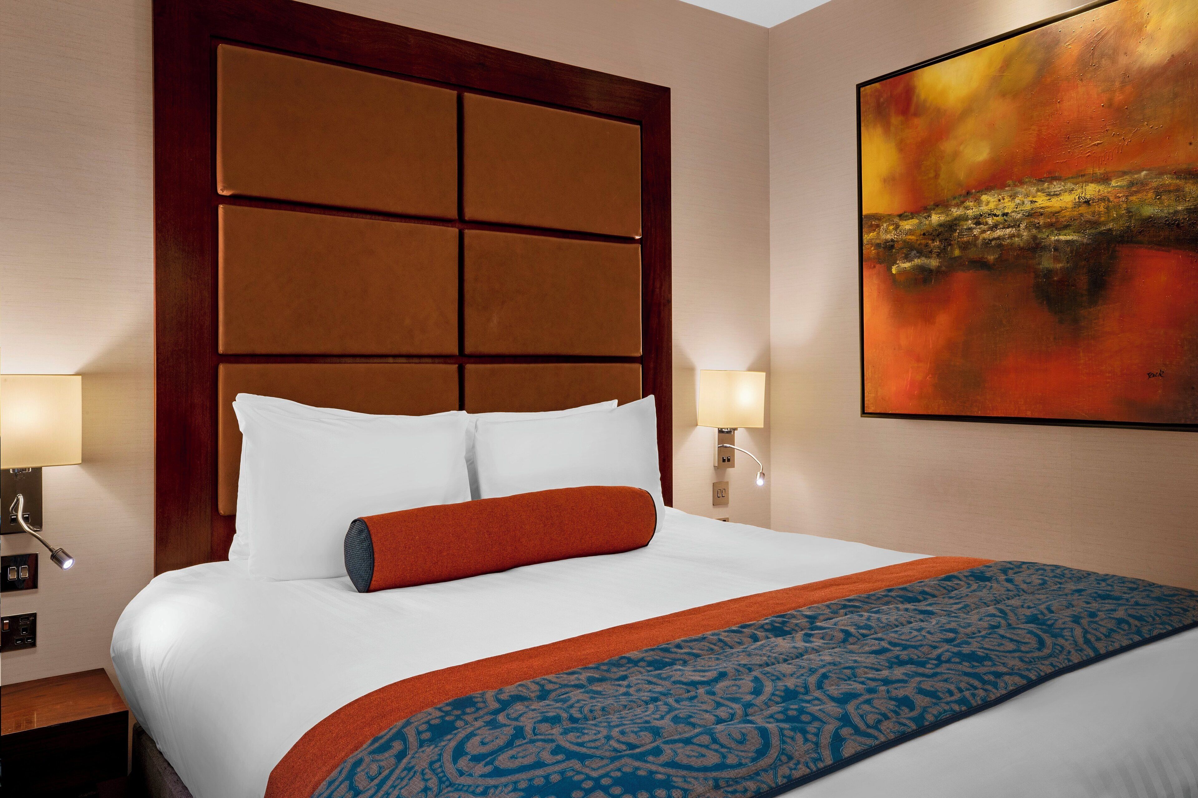 A hotel room with a large bed, two bedside lamps, a headboard with a panel design, and a painting on the wall.