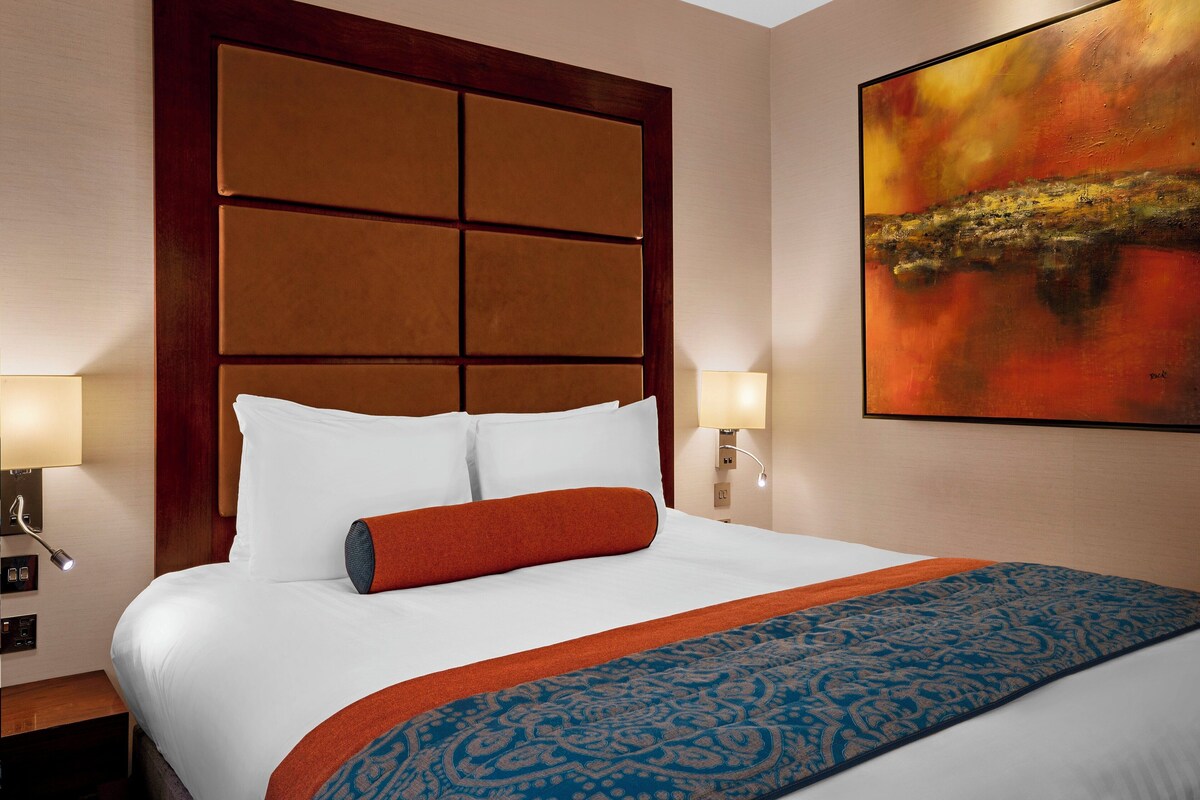 A hotel room with a large bed, two bedside lamps, a headboard with a panel design, and a painting on the wall.