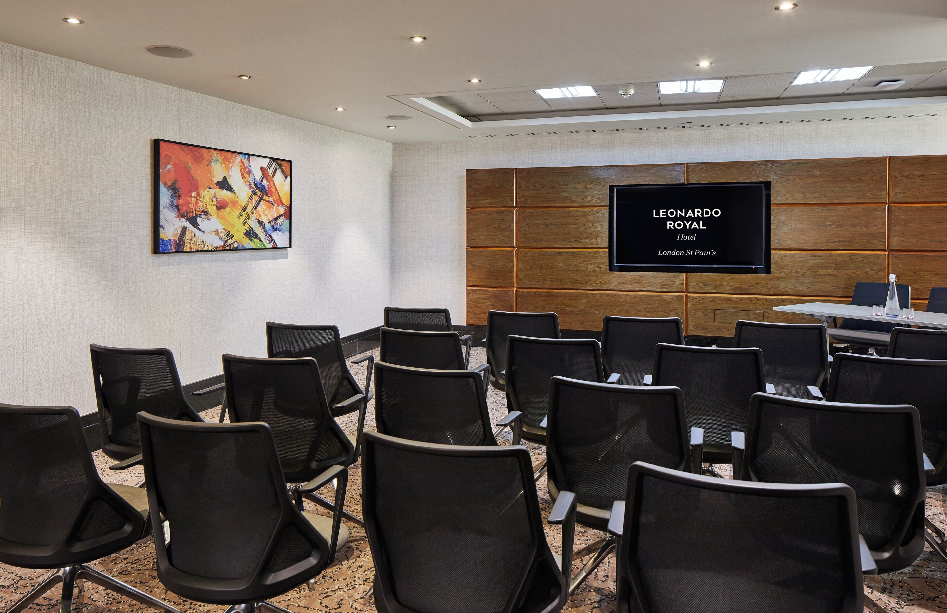 Ein Konferenzraum mit schwarzen Stühlen, die in Reihen angeordnet sind, einem großen Flachbildfernseher an der Wand und einem farbenfrohen, abstrakten Gemälde darüber.