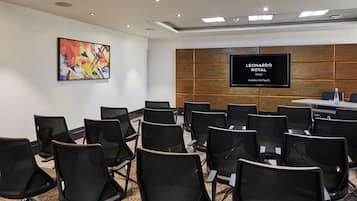 Ein Konferenzraum mit schwarzen Stühlen, die in Reihen angeordnet sind, einem großen Flachbildfernseher an der Wand und einem farbenfrohen, abstrakten Gemälde darüber.