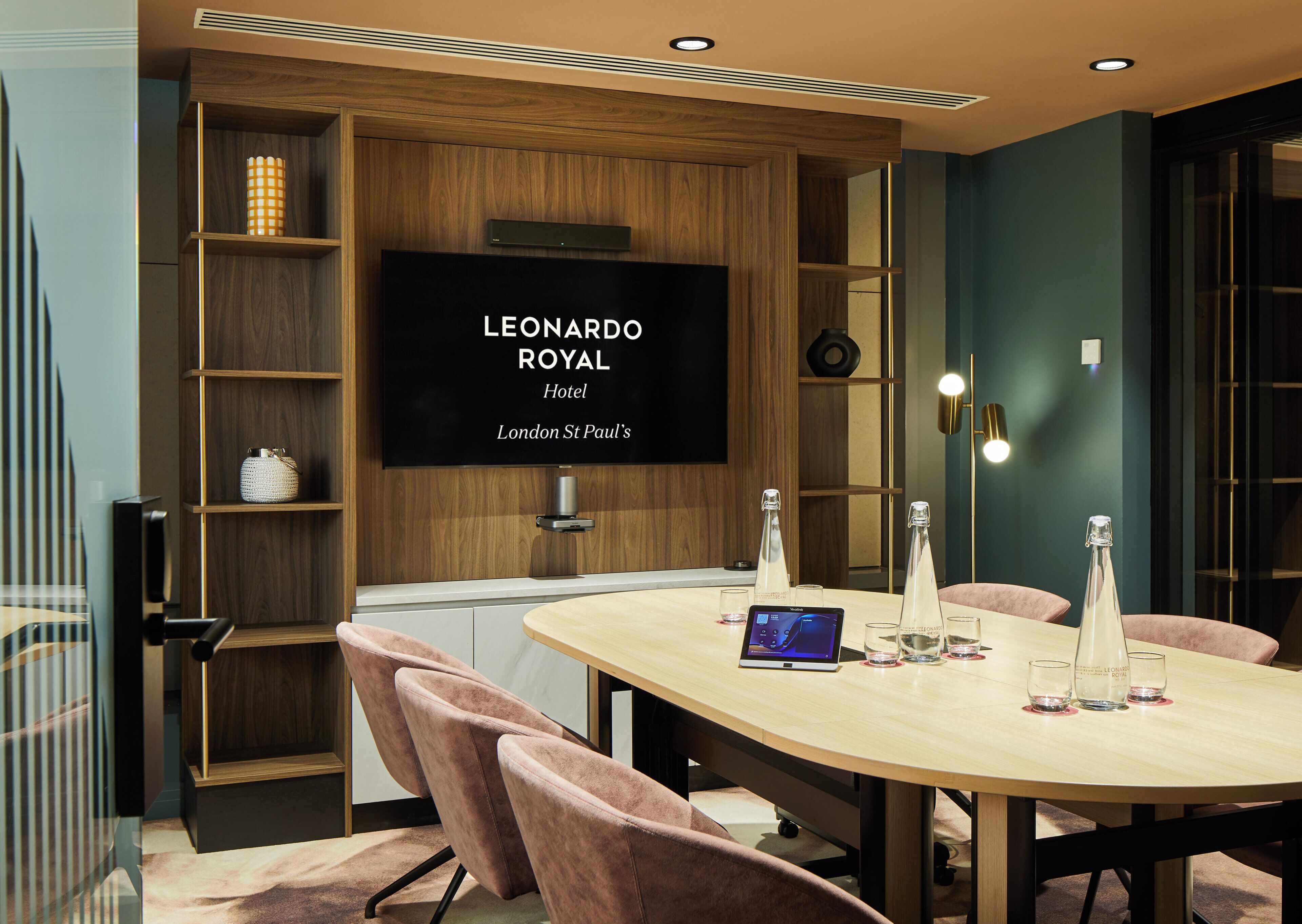 A modern hotel room with a round table, pink chairs, a flat-screen TV, and a bookshelf.