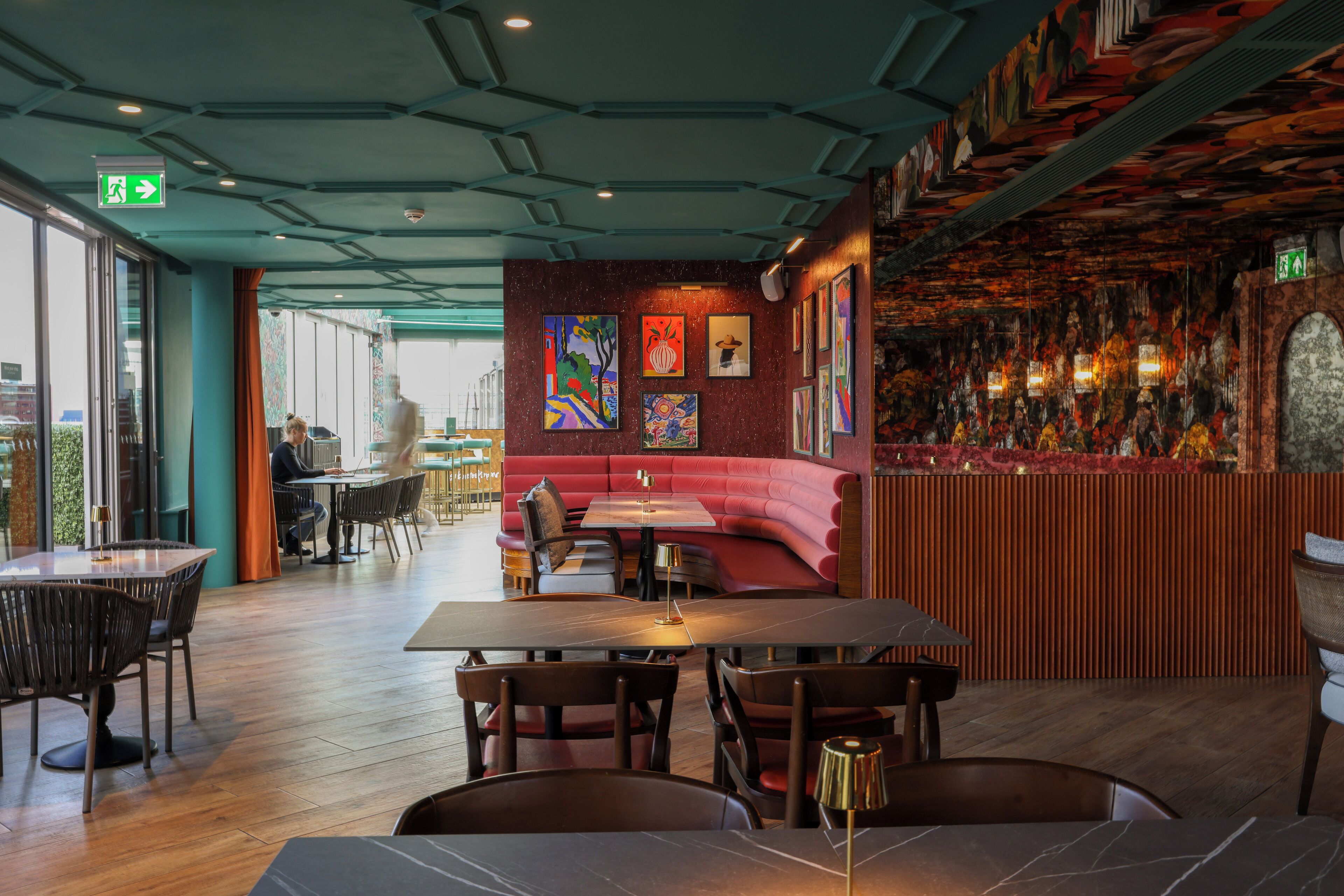 L’intérieur d’un restaurant est aménagé avec des tables et des chaises en bois, un coin salon avec des banquettes rouges et des œuvres d’art accrochées aux murs.