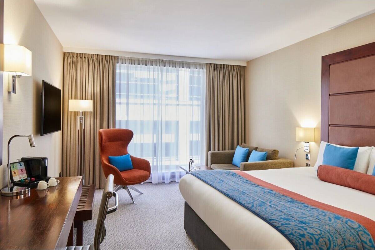 Ein Hotelzimmer mit Bett, Schreibtisch mit Kaffeemaschine, Stuhl, Sofa und Fernseher.