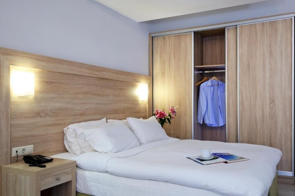Una habitación de hotel con cabecera de madera, una mesita de noche con teléfono, cama con ropa blanca, armario con una prenda colgada y un jarrón con flores.