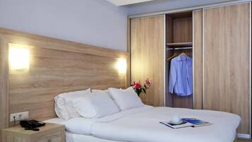 Una habitación de hotel con cabecera de madera, una mesita de noche con teléfono, cama con ropa blanca, armario con una prenda colgada y un jarrón con flores.