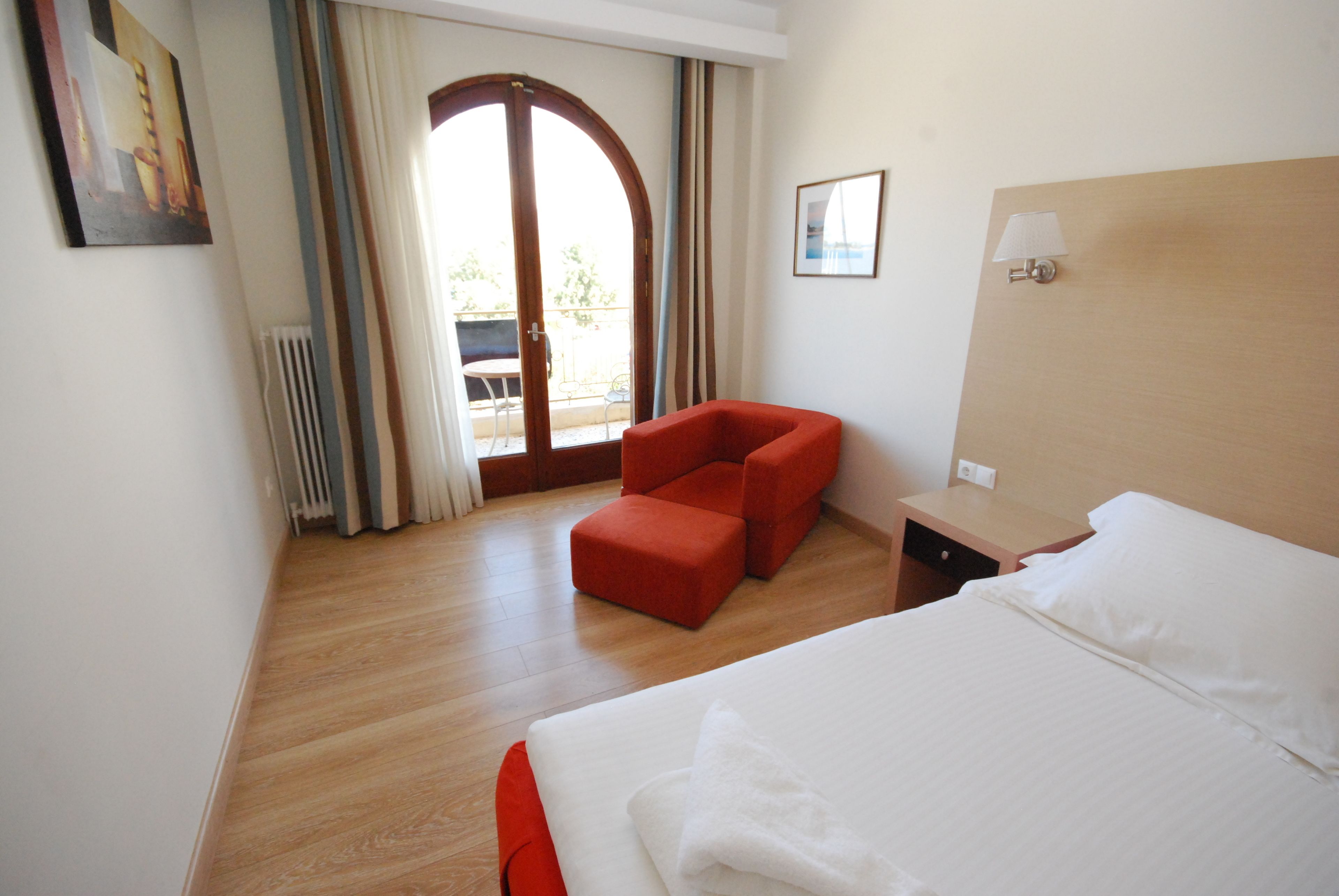 Une chambre d’hôtel avec un grand lit, un fauteuil rouge, une petite table et une vue sur l’extérieur grâce à une porte vitrée.