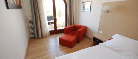Quarto de hotel com uma cama grande, uma poltrona vermelha, uma mesa pequena e vista para o exterior através de uma porta de vidro.
