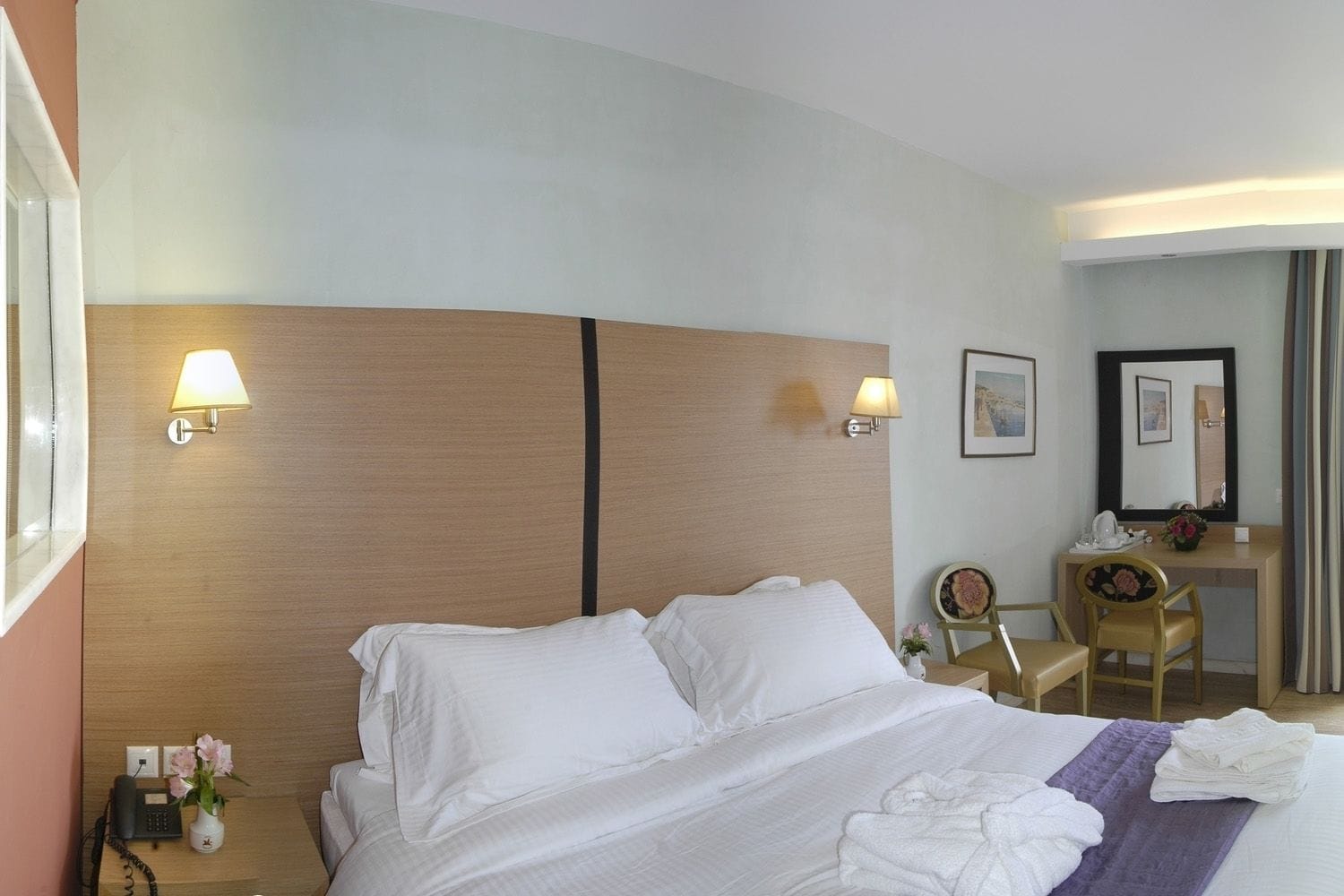 A hotel room with a large bed, bedside lamps, a desk with a chair, a phone, and a vase with flowers.