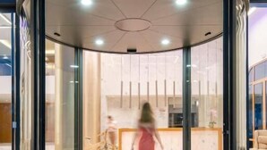 Une porte tournante à panneaux de verre, donnant sur un espace d’accueil où se trouvent un vélo et une personne portant une robe rouge.