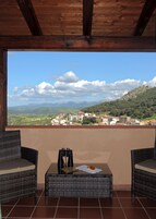 Un balcón con mobiliario de mimbre, binoculares y vistas a un pueblo y montañas.