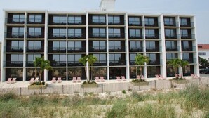 A multi-story hotel with balconies, a sandy beach area with lounge chairs, and palm trees.